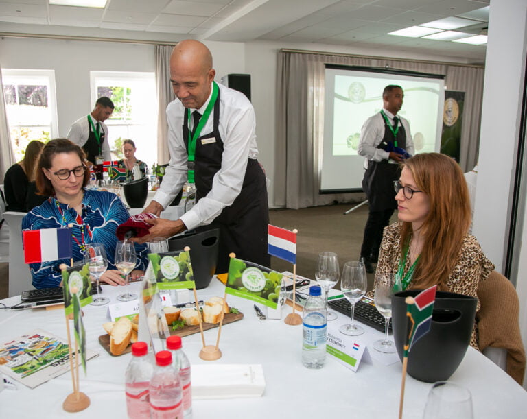 Judges being served white wine. There are judges' countries flags on the table on the table. A france flag on the left and a Nederlands flag on the right